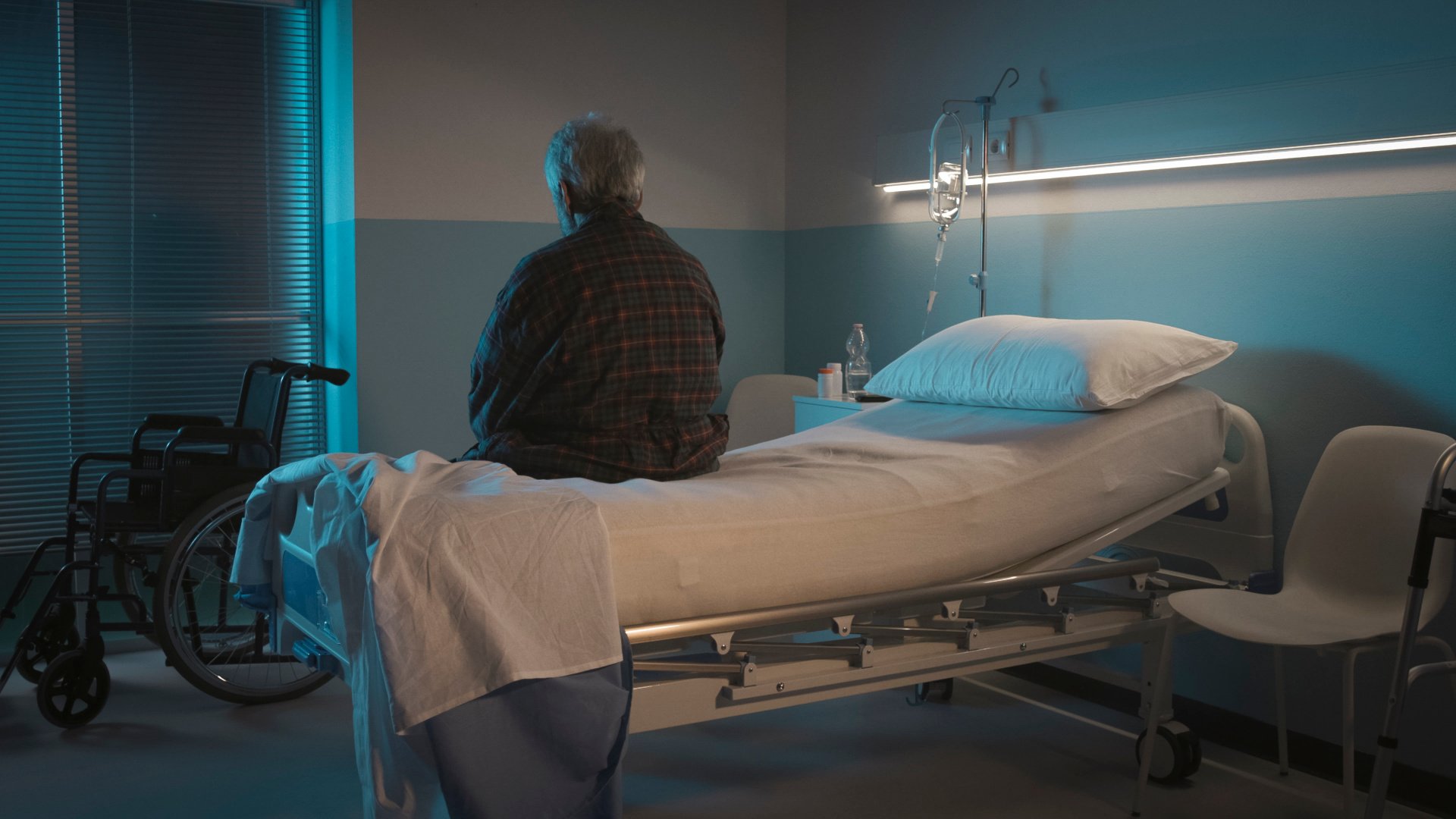 ryggen til en eldre pasient sitter på en sykehusseng og ser ned på gulvet i et mørk sykehus rom